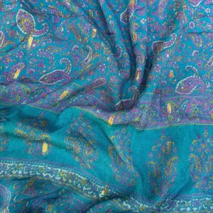 May include: A close-up of a teal fabric with a paisley pattern in shades of purple, yellow, and gold. The fabric is draped, showing folds and texture. The paisley design is repeated across the surface.