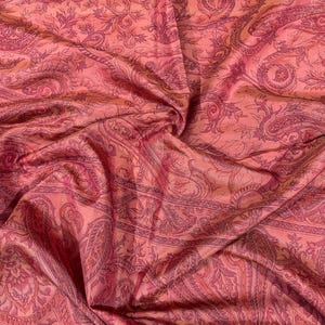 May include: A close-up of a fabric with a paisley pattern in shades of pink and red. The fabric is draped, showing folds and texture. The design features intricate floral and curved elements.