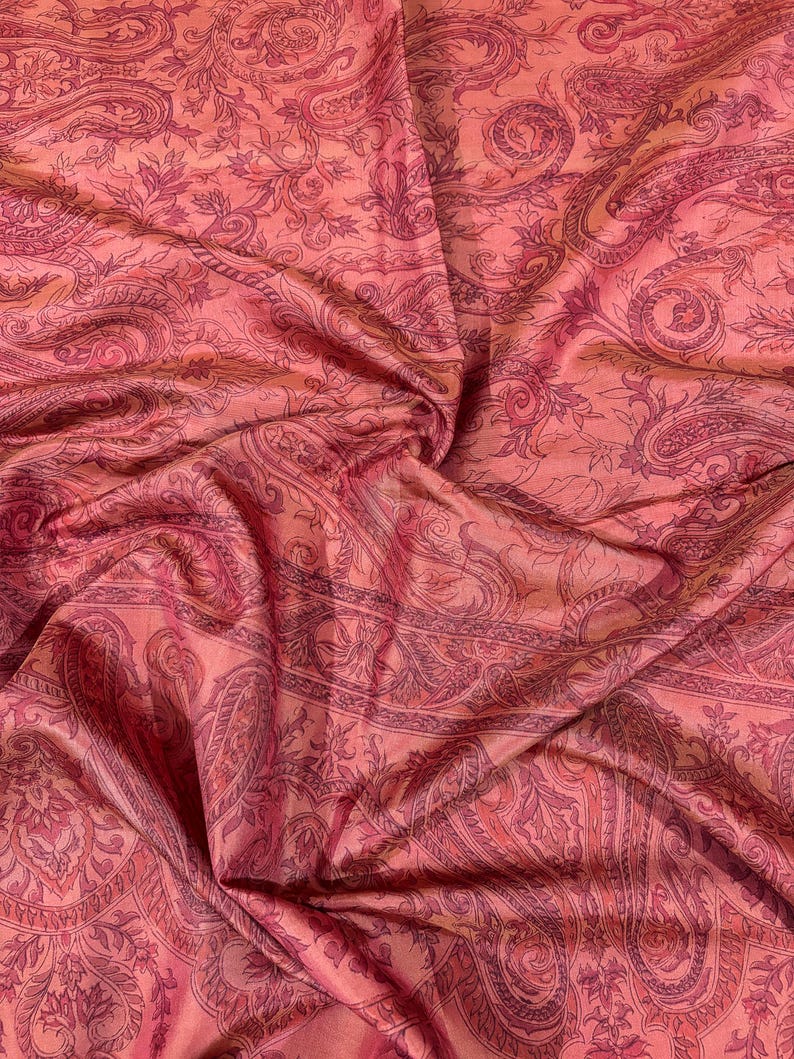 May include: A close-up of a fabric with a paisley pattern in shades of pink and red. The fabric is draped, showing folds and texture. The design features intricate floral and curved elements.