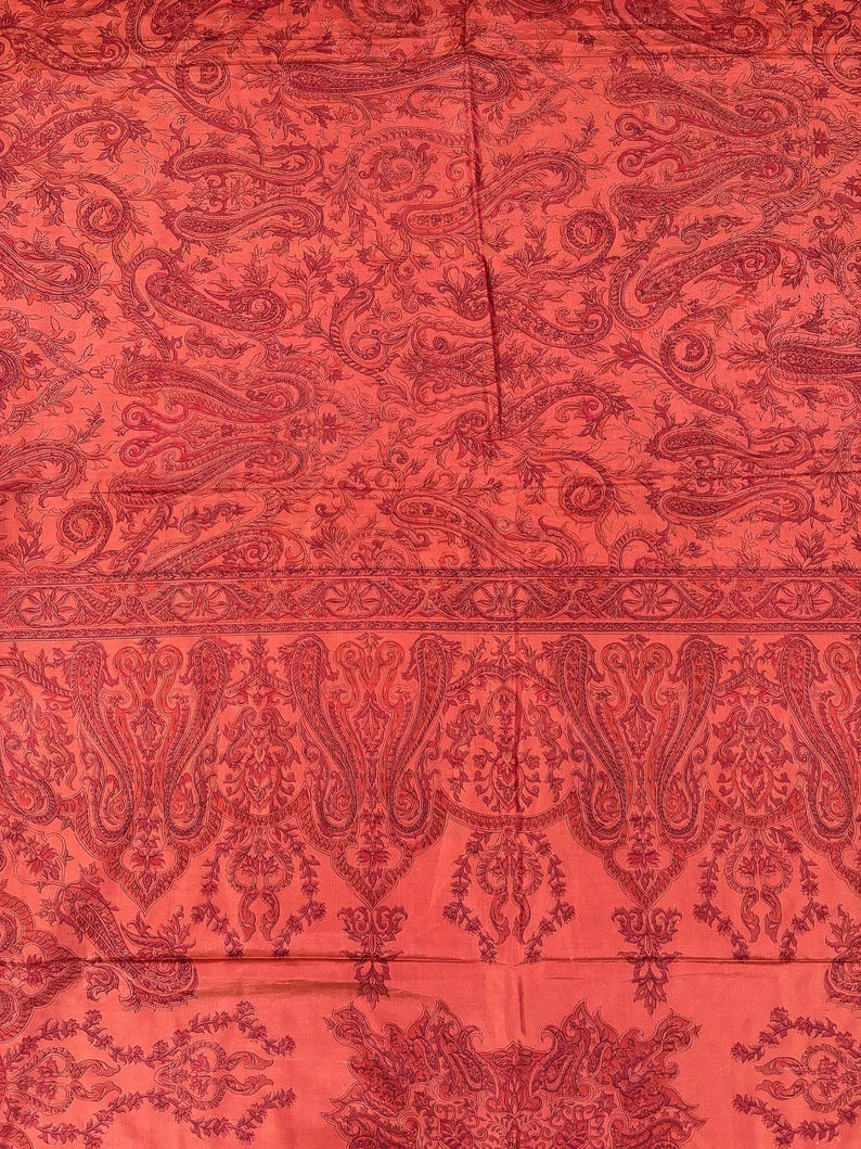 May include: A close-up of a red textile with a paisley pattern. The fabric features a repeating design of curved, teardrop-shaped motifs in a darker shade of red. The overall effect is a rich, detailed, and ornate design.