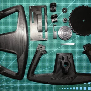 May include: A set of black 3D printed parts for a steering wheel. The parts include a steering wheel rim, two steering wheel grips, a gear shifter, and a button panel. The parts are laid out on a green cutting mat.