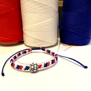 May include: A red, white, and blue friendship bracelet with a white soccer ball charm. The bracelet is made with a braided cord.
