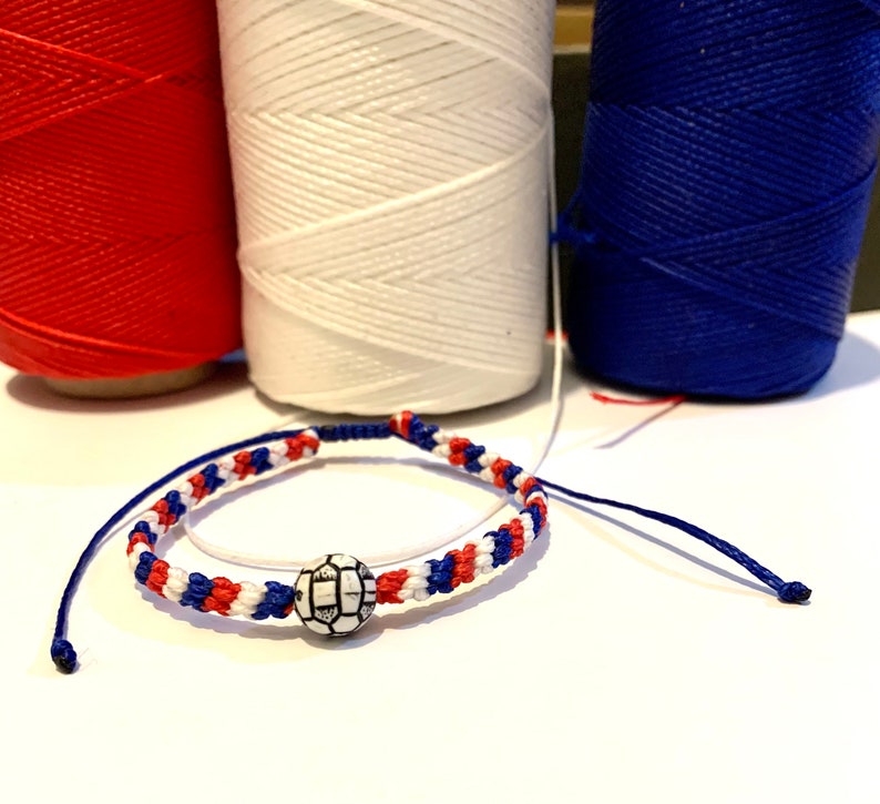 May include: A red, white, and blue friendship bracelet with a white soccer ball charm. The bracelet is made with a braided cord.