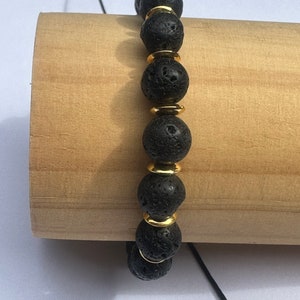 Può includere: Un braccialetto in pietra lavica nera con dettagli dorati. Il braccialetto è composto da perline rotonde e infilate su un cordoncino nero.