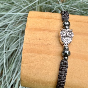 May include: A dark brown macrame bracelet featuring a silver owl charm with intricate detailing. The bracelet includes dark gray beads and is displayed on a wooden surface against a backdrop of green foliage.