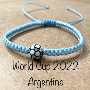 以下が含まれることがあります： サッカーボールのチャームが付いた青と白の友情ブレスレット。ブレスレットには、茶色の背景に「ワールドカップ2022 アルゼンチン」の文字が書かれています。