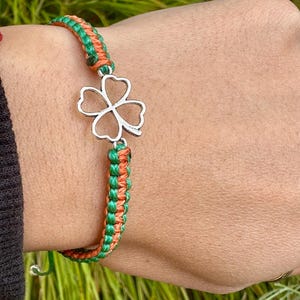 May include: A green and orange braided bracelet with a silver four-leaf clover charm. The bracelet is made of woven cord and is displayed on a person's wrist.