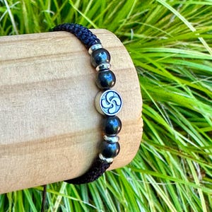 May include: Black beaded bracelet with silver accents and a central silver charm featuring a Basque Lauburu symbol. The bracelet is made of black cord and dark beads. The bracelet is displayed on a wooden surface with green grass in the background.