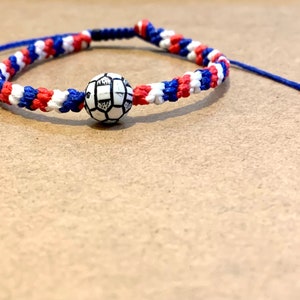 May include: A blue, white, and red braided bracelet with a white and black soccer ball charm.