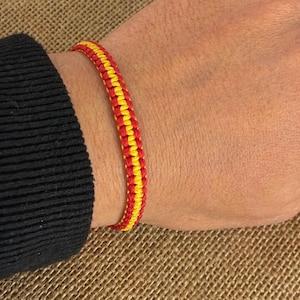 Puede incluir: Una pulsera tejida en rojo y amarillo, usada en una muñeca. La pulsera está hecha de hilos tejidos apretadamente, con los colores alternando en un patrón repetitivo. La muñeca es de piel clara y se ve una manga negra.