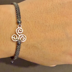 May include: A grey braided bracelet with a silver Celtic triskele charm. The charm has a three-spiral design. The bracelet is on a wrist.