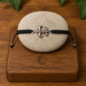 Può includere: Un bracciale in cordino nero con un ciondolo centrale a forma di giglio in tonalità argento. Il bracciale presenta estremità intrecciate regolabili rifinite con piccole perline argentate. Il design è centrato su una pietra ovale chiara.