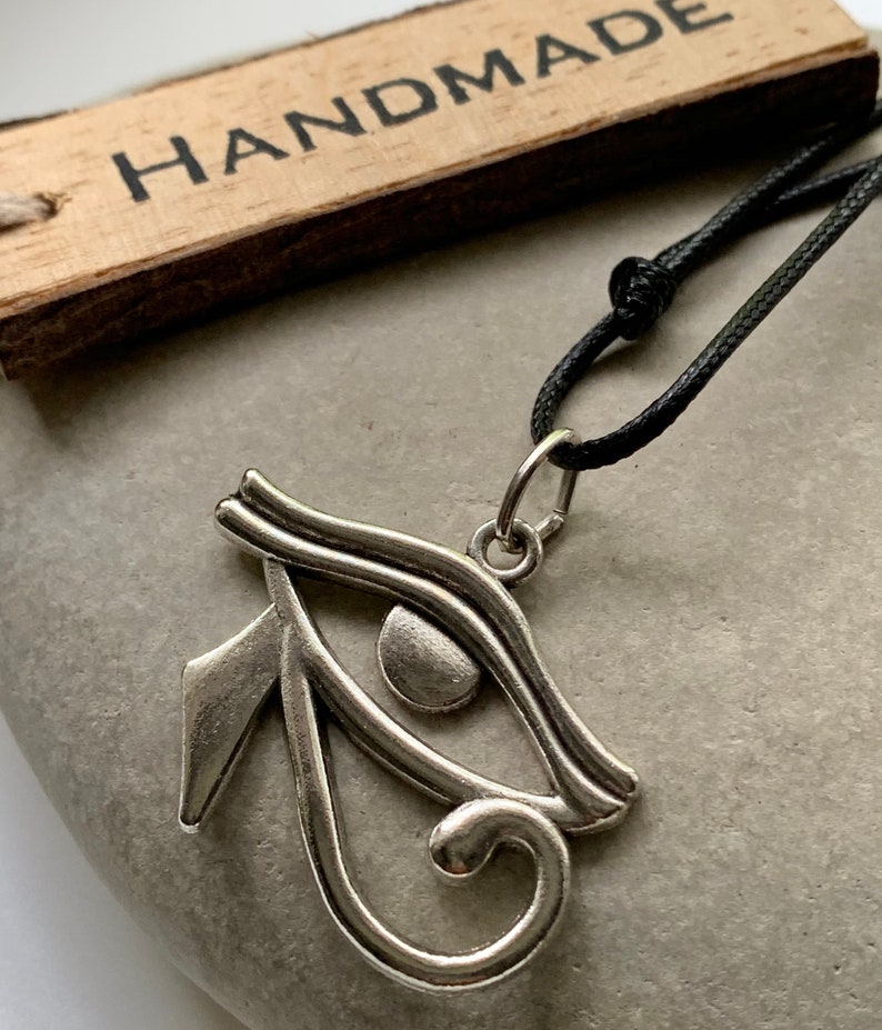 May include: A silver Eye of Horus pendant necklace on a black cord. The pendant is a detailed representation of the ancient Egyptian symbol. The necklace is handmade.