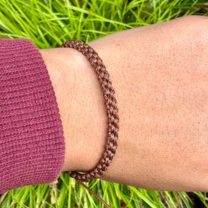 Puede incluir: Una pulsera de macramé marrón se usa en la muñeca, mostrando un diseño tejido. La pulsera está hecha de un material marrón claro y se asegura con un cordón fino. El fondo presenta hierba verde y una manga granate.