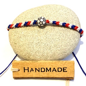 May include: Handmade friendship bracelet with red, white, and blue braided cords and a black and white soccer ball charm. The bracelet is displayed on a gray stone with a wooden tag that says "HANDMADE".