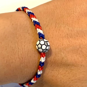 May include: A red, white, and blue braided bracelet with a black and white soccer ball charm.