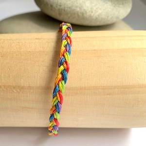 Könnte beinhalten: Ein geflochtenes Armband in Regenbogenfarben, darunter Rot, Orange, Gelb, Grün, Blau und Lila. Das Armband besteht aus gewebten Fäden und wird auf einer Holzoberfläche mit einem grauen Stein im Hintergrund präsentiert.