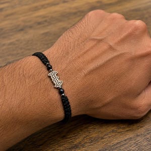 以下が含まれることがあります： 黒い編み込みコードのブレスレット。中央には穴あきテクスチャの銀色のコウモリ型チャームがあり、その両側に小さな暗いメタリックビーズが配置され、編み込みノットのデザインで手首に固定されています。