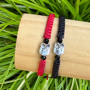 Puede incluir: Dos pulseras trenzadas con dijes de búho. Una pulsera es roja y la otra es negra.