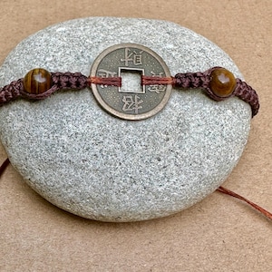 May include: A brown macrame bracelet featuring a bronze coin with Chinese characters and two tiger eye beads. The bracelet is displayed on a gray stone, showcasing the intricate details of the design.