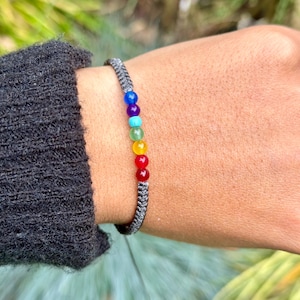 Puede incluir: Una pulsera de macramé gris con siete cuentas de chakra coloridas. Las cuentas están dispuestas en una fila, mostrando los colores del arcoíris. La pulsera se lleva en una muñeca, con una manga gris oscura visible.