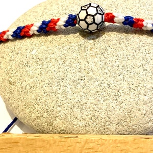 May include: A red, white, and blue braided bracelet with a black and white soccer ball charm. The bracelet is handmade and is displayed on a wooden surface with the word "HANDMADE" written on it.