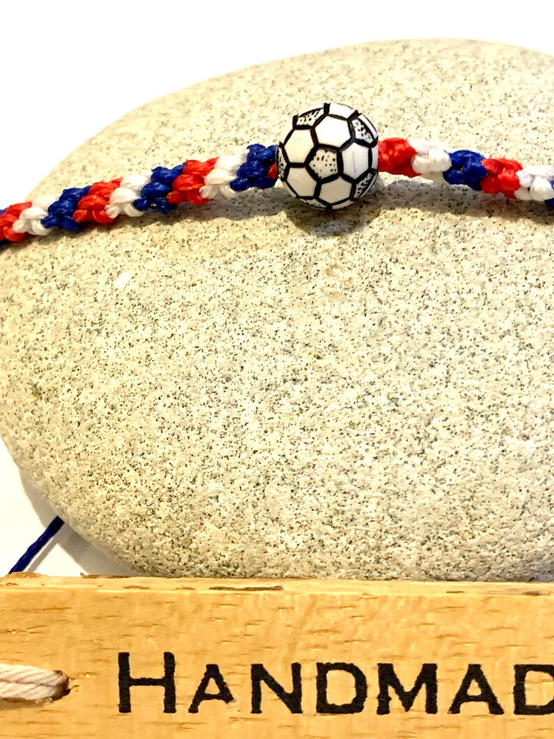 May include: A red, white, and blue braided bracelet with a black and white soccer ball charm. The bracelet is handmade and is displayed on a wooden surface with the word "HANDMADE" written on it.