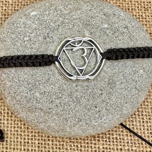Op de afbeelding: Zilverkleurige chakra-symboolarmband met een zwart geweven koord. De armband heeft een cirkelvormige hanger met een geometrisch ontwerp en het nummer 3 in het midden. De armband wordt getoond op een grijze steen.