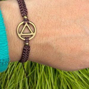 May include: A brown macrame bracelet with a gold-colored circular charm featuring a triangle symbol. The bracelet is on a wrist, with a green background of grass.