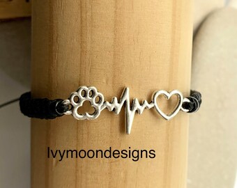 May include: Black braided bracelet with a silver paw print, heartbeat, and heart charm. The bracelet is on a wooden surface. The text "ivymoondesigns" is visible below the bracelet.