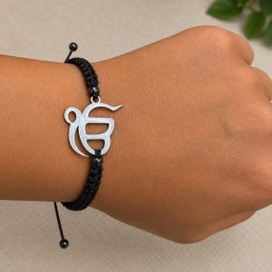 Könnte beinhalten: Ein Armband aus schwarzer Kordel mit einem silberfarbenen Metallanhänger in Form des Ik Onkar-Symbols. Das Armband besteht aus einer geflochtenen schwarzen Kordel und enthält kleine schwarze Perlen auf beiden Seiten des zentralen Metallemblems.