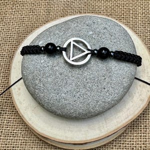 以下が含まれることがあります： 黒いマクラメブレスレット。シルバーの円の中に三角形のチャームと黒いビーズが付いています。ブレスレットは、ジュートの背景に対して、木の輪切りの上に置かれた灰色の石の上に展示されています。