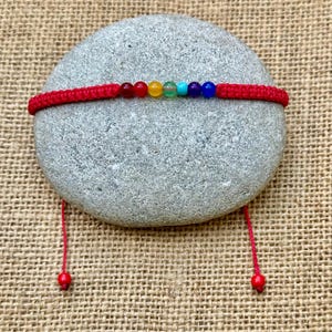 Puede incluir: Una pulsera de macramé roja con siete cuentas de colores que representan los chakras. La pulsera se muestra sobre una piedra gris sobre un fondo texturizado y beige. La pulsera tiene cordones rojos ajustables con pequeñas cuentas rojas.