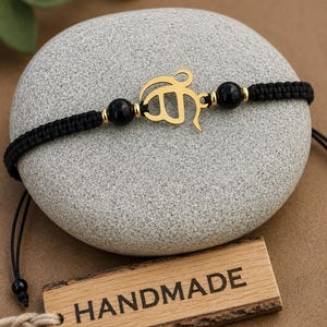 May include: A black cord bracelet featuring a gold-coloured metal Khanda symbol at the centre, flanked by two round black beads and small gold-coloured spacers. The bracelet has an adjustable woven cord design and is displayed on a smooth grey stone.