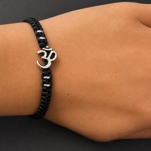 Può includere: Un bracciale in corda intrecciata nera con un ciondolo centrale a forma di simbolo Om color argento. Il bracciale è arricchito da piccole perline metalliche cilindriche color argento integrate nella corda nera intrecciata e resistente.