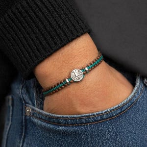 Puede incluir: Una pulsera de macramé con un dije plateado de árbol de la vida. La pulsera presenta hilos marrones y turquesas, con cuentas plateadas. La pulsera se usa en una muñeca, con un suéter negro y jeans azules en el fondo.