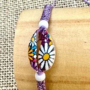 May include: A purple and white braided bracelet with a shell charm featuring a floral pattern. The shell is decorated with colourful daisies.