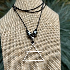 Könnte beinhalten: Eine schwarze Kordelkette mit einem silbernen Dreiecksanhänger. Die Halskette hat schwarze Perlen und eine dekorative silberne Perle. Der Anhänger hat eine geometrische Form, und die Halskette wird auf einer beigen Oberfläche präsentiert.
