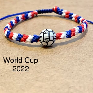 May include: A red, white, and blue braided friendship bracelet with a white soccer ball charm. The bracelet has the text "World Cup 2022" printed below it.