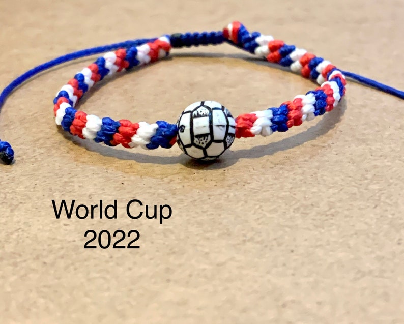 May include: A red, white, and blue braided friendship bracelet with a white soccer ball charm. The bracelet has the text "World Cup 2022" printed below it.