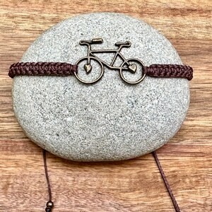 May include: A brown braided bracelet with a metal bicycle charm. The bicycle charm has two hearts on the wheels.