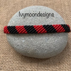 May include: Black and red friendship bracelet with a checkered pattern. The bracelet is on a grey rock.