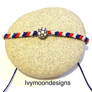 May include: A red, white, and blue friendship bracelet with a white soccer ball charm. The bracelet is on a gray rock.