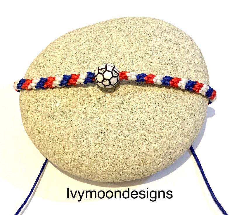 May include: A red, white, and blue friendship bracelet with a white soccer ball charm. The bracelet is on a gray rock.