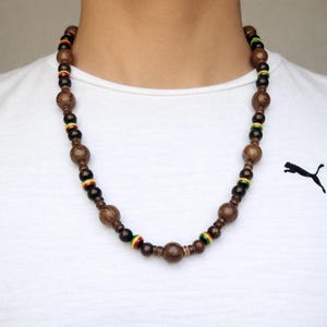 Könnte beinhalten: Eine Holzkette mit Rastafari-Farbschema. Die Halskette besteht aus braunen Holzperlen in verschiedenen Größen, die mit kleineren Perlen in Rot, Gelb und Grün durchsetzt sind. Die Halskette wird auf einem weißen T-Shirt getragen.