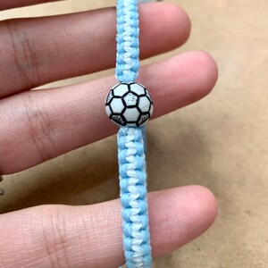 以下が含まれることがあります： 白と水色の友情ブレスレットに、白黒のサッカーボールのチャームが付いている