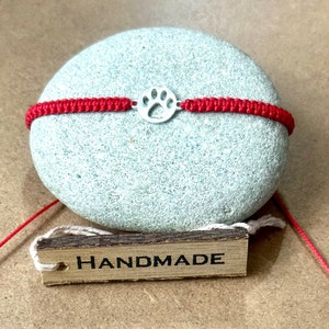 May include: Red string bracelet with a silver paw print charm. The bracelet is handmade and has a wooden tag with the word "HANDMADE" written on it.