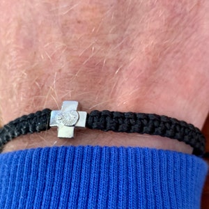 May include: A black braided bracelet with a silver cross charm. The bracelet is worn on a person's wrist.