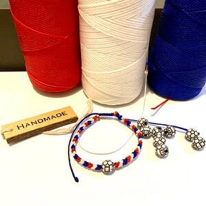 May include: Handmade bracelet with red, white, and blue string and soccer ball beads. The bracelet is on a white surface with a wooden tag that says "HANDMADE".