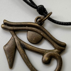 May include: A bronze-colored Eye of Horus pendant on a black cord necklace. The pendant is detailed with intricate lines and a circular eye.